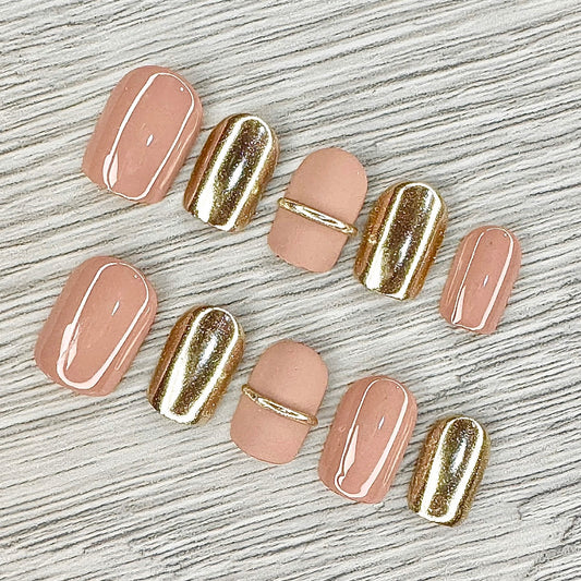 Elegant press-on nails featuring a combination of chic nude and luxurious gold accents, beautifully displayed on a textured background. The set includes various nail sizes with a mix of glossy and matte finishes, perfect for adding a touch of glamour to any look